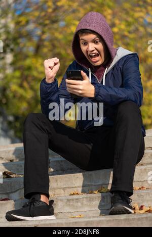 Stupito adolescente felice che guarda lo schermo del cellulare seduto sulle scale all'aperto. Ragazzo adolescente scioccato dalla sua fortuna Foto Stock