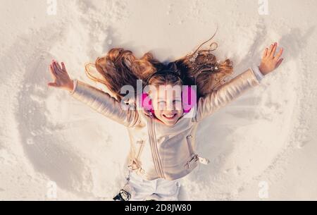 Capretto divertente che fa l'angelo della neve. Bambina che gioca e fa un angelo di neve nella neve. Vista dall'alto. Foto Stock