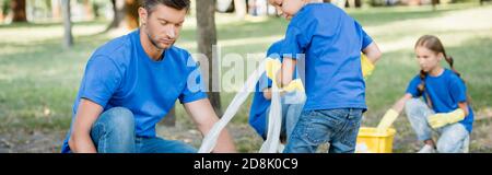 padre e figlio che raccolgono rifiuti di plastica nella foresta vicino a madre e figlia su sfondo offuscato, concetto di ecologia, banner Foto Stock