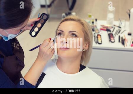 Ritratto di affascinante bella donna matura trendy che ha fatto il trucco E un artista MUA che applica l'ombra degli occhi con il pennello i suoi occhi hanno una tavolozza di colori Foto Stock