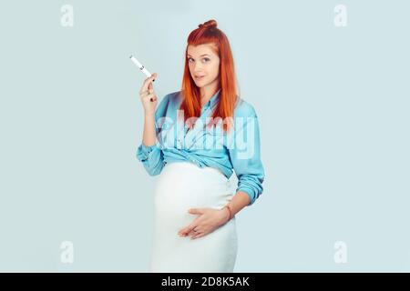 Giovane rosso bella donna che tocca il ventre incinta e tiene la sigaretta elettronica e-sigaretta guardando la macchina fotografica isolata su sfondo blu. Misto Foto Stock