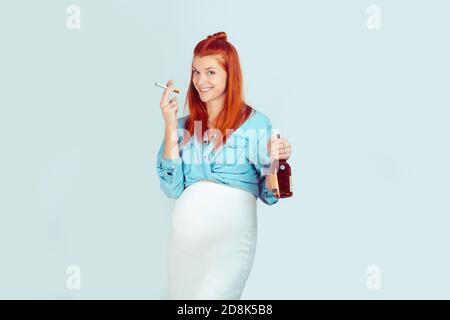 Giovane ragazza incauta con i capelli di zenzero in attesa per il bambino e in piedi con sigaretta e alcool sorridente felice su sfondo azzurro chiaro. Modello di gara misto Foto Stock