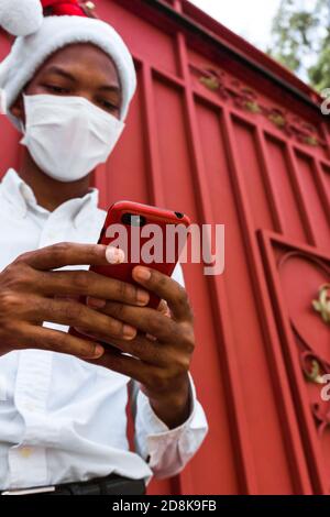 Uomo afro-americano che tiene il suo telefono con custodia rossa che indossa Babbo Natale cappello in piedi contro un cancello rosso Foto Stock