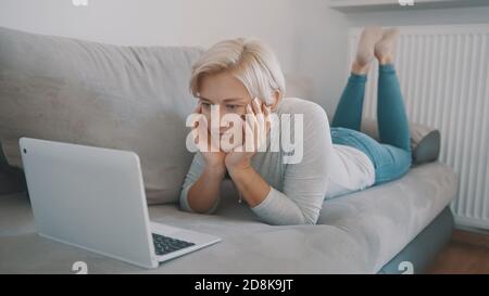 Giovane donna tourghtful che chiacchiera con i frends sui social media usando il laptop sul suo divano. Foto di alta qualità Foto Stock