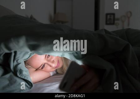 Giovane donna allegra sdraiata a letto sotto la coperta utilizzando il telefono cellulare a tarda notte, non può dormire. Insonnia, nomofobia, concetto di dipendenza da Internet Foto Stock