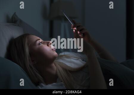 Giovane donna spossata sonnolenta sdraiata a letto usando il telefono cellulare, non può dormire. Insonnia, nomofobia, disturbi del sonno concetto Foto Stock