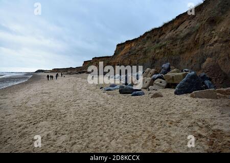 Una passeggiata in famiglia lungo la spiaggia di Happisburgh, accanto alle scogliere sul mare che mostrano l'erosione costiera a Norfolk, Inghilterra. Grandi rocce si ergono lungo la scogliera. Foto Stock