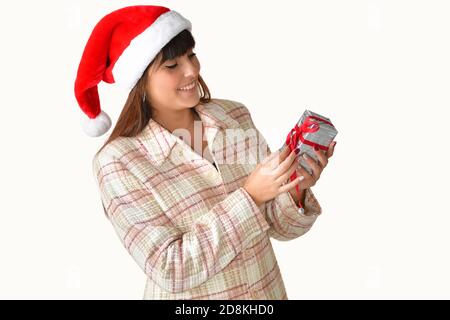 Bella giovane donna che indossa un cappello di Santa guardando una scatola regalo in argento con un nastro rosso su uno sfondo chiaro. Natale e regali concetto. Foto Stock