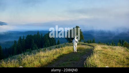 Vista posteriore del viaggiatore di spazio a piedi sulla strada di montagna con la chitarra. Chitarrista cosmonauta in tuta spaziale bianca camminando lungo il sentiero erboso attraverso il prato con colline foggose e cielo sullo sfondo. Foto Stock
