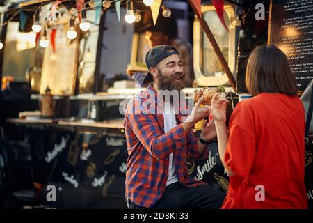 una coppia urbana si diverte di fronte al fast food mobile restaurant, mangiando un panino insieme, divertendosi, ridendo Foto Stock