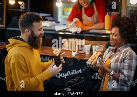 donna dipendente in fast food servizio sorridente, guardando carina multietnica coppia in amore mangiare panini Foto Stock