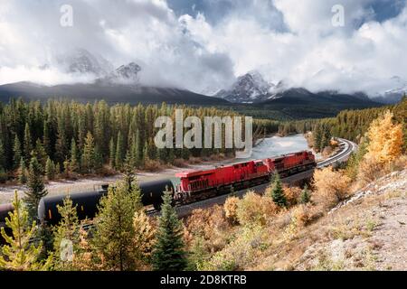 Treno lungo ferroviario rosso che passa attraverso la valle autunnale con la montagna a Morant's Curve, Calgary, Canada Foto Stock