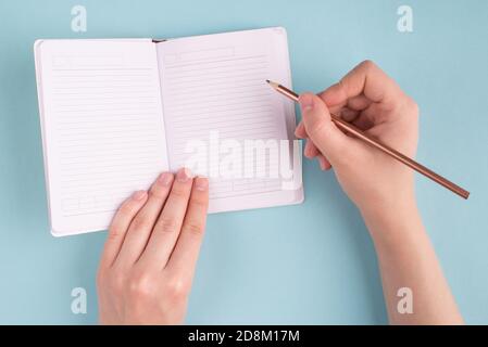 Sopra la vista ravvicinata della foto delle mani femminili della ragazza tenendo la matita che inizia a fare i compiti in copia libro isolato su sfondo blu di colore pastello Foto Stock