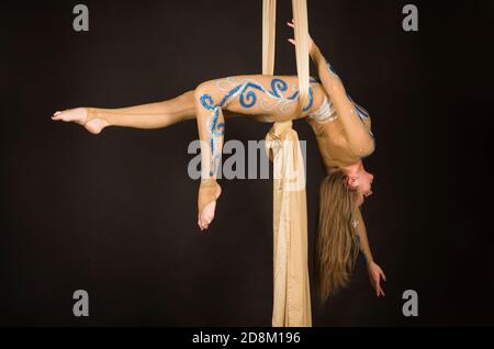 Una donna snella in una tuta beige esegue ginnastica ed esercizi circensi sulla seta dorata. Riprese in studio su sfondo scuro, immagini isolate. Foto Stock