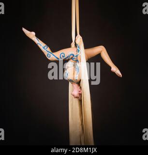Una donna snella in una tuta beige esegue ginnastica ed esercizi circensi sulla seta dorata. Riprese in studio su sfondo scuro, immagini isolate. Foto Stock