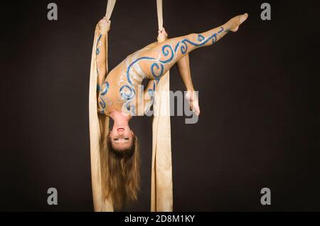 Una donna snella in una tuta beige esegue ginnastica ed esercizi circensi sulla seta dorata. Riprese in studio su sfondo scuro, immagini isolate. Foto Stock