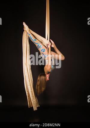 Una donna snella in una tuta beige esegue ginnastica ed esercizi circensi sulla seta dorata. Riprese in studio su sfondo scuro, immagini isolate. Foto Stock