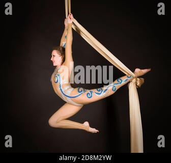 Una donna snella in una tuta beige esegue ginnastica ed esercizi circensi sulla seta dorata. Riprese in studio su sfondo scuro, immagini isolate. Foto Stock