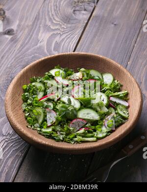 Insalata vitaminica di erbe selvatiche con cetriolo, rafano e cipolle verdi in una ciotola di legno su fondo di legno. Cibo sano disintossicante Foto Stock