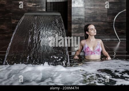 Bella giovane femmina sottile in bikini seduta in acqua gorgogliante di piscina con cascata e godendo sessione di idroterapia in centro benessere Foto Stock