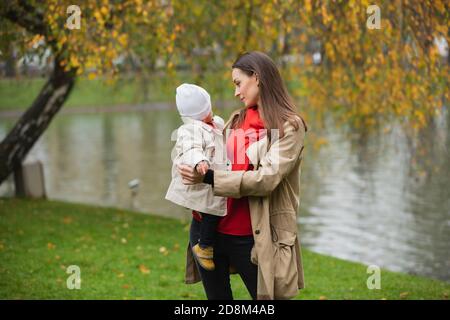 Mamma e il suo bambino camminano nel parco autunnale con la sua bambina tra le braccia, un lago dietro di loro Foto Stock