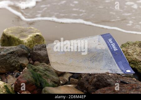 La protezione del viso è stata scartata e lavata sulla spiaggia causando un problema ambientale con la plastica dei rifiuti durante la pandemia. Foto Stock