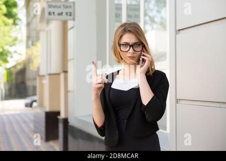 Giovane donna rigida con smartphone che non mostra, non, attenzione con il gesto della mano delle dita all'esterno vicino al negozio della casa d'ufficio. Parlare con i bambini, genitori Foto Stock