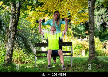 Un uomo alla fine degli anni sessanta solleva i pesi all'aperto con l'aiuto di una giovane personal trainer femminile. Foto Stock