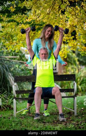 Un uomo alla fine degli anni sessanta solleva i pesi all'aperto con l'aiuto di una giovane personal trainer femminile. Foto Stock