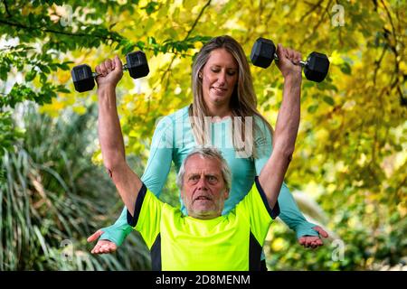 Un uomo alla fine degli anni sessanta solleva i pesi all'aperto con l'aiuto di una giovane personal trainer femminile. Foto Stock