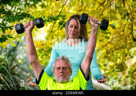 Un uomo alla fine degli anni sessanta solleva i pesi all'aperto con l'aiuto di una giovane personal trainer femminile. Foto Stock