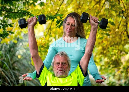 Un uomo alla fine degli anni sessanta solleva i pesi all'aperto con l'aiuto di una giovane personal trainer femminile. Foto Stock