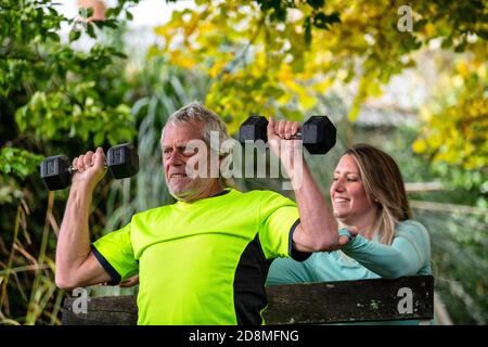 Un uomo alla fine degli anni sessanta solleva i pesi all'aperto con l'aiuto di una giovane personal trainer femminile. Foto Stock