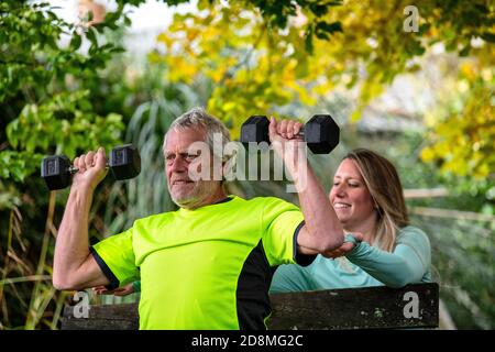 Un uomo alla fine degli anni sessanta solleva i pesi all'aperto con l'aiuto di una giovane personal trainer femminile. Foto Stock