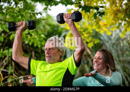 Un uomo alla fine degli anni sessanta solleva i pesi all'aperto con l'aiuto di una giovane personal trainer femminile. Foto Stock