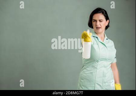 Giovane donna che indossa una camicia verde e guanti gialli, tenendo la soluzione detergente per vetri in mani isolate su sfondo verde Foto Stock