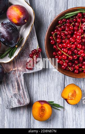 Frutta fresca di prugne dolce intera e tagliata in piatto con foglie di rosmarino sul vecchio tagliere con bacche di ribes rosso in una ciotola di legno Foto Stock