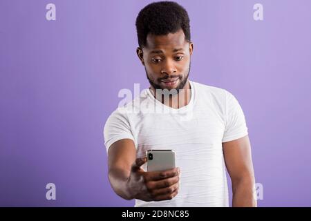 Studio shot di allegro sorridente african american barba lunga giovane azienda cellulare, tenendo selfie ritratto, rendendo la chiamata video, indossando vestiti di bianco Foto Stock