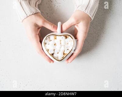 Le mani femminili tengono la tazza bianca di ceramica sotto forma di cuore con la bevanda calda e il marshmallows su sfondo chiaro. Vista dall'alto. Spazio di copia Foto Stock