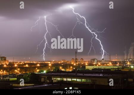 Potente lampo che colpisce la città di notte su un cielo grigio scuro. Un forte fulmine colpisce il terreno, illuminando l'a industriale Foto Stock