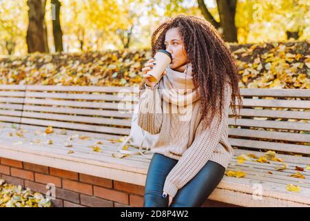 Bella donna afro capelli indossando maglia maglione e sciarpa sedersi sulla panchina in autunno parco in giornata di sole con caffè. Foto Stock