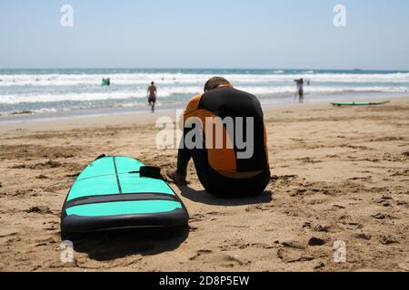 Surfer principiante in un costume da bagno è seduto sulla sabbia bagnata sull'oceano. La sua testa è piegata su mani piegate. Accanto c'è una tavola da surf. Surfista Foto Stock