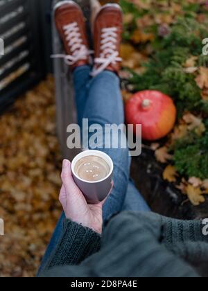 Donna in felpa verde e jeans blu che tengono una tazza di caffè con le parole - rimanere a casa, stare al sicuro. Decorazione d'autunno, foglie cadute. Vista dall'alto. Foto Stock