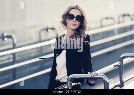 Moda giovane bionda business donna appoggiata sulla ringhiera elegante modello femminile indossando giacca nera e occhiali da sole Foto Stock