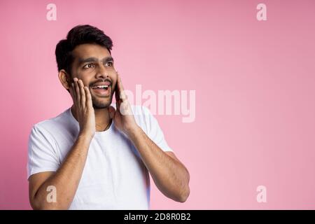 Ritratto di felice sorpreso piacevolmente uomo in t shirt bianca, toccante guance con palme, guardando da parte con espressione stupita, isolato su sfondo rosa Foto Stock