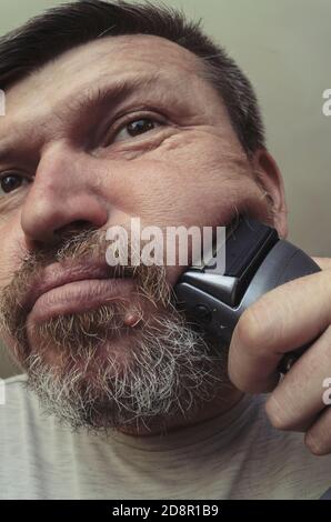 Un uomo adulto si rasa con un rasoio elettrico. Ritratto insolito di un uomo bearded con capelli grigi. Grimaces davanti alla fotocamera Foto Stock