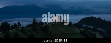 Jamnik, Slovenia - Vista panoramica dell'ora blu a Jamnik con la chiesa illuminata di San Primoz in cima a una nebbia alba estiva. Alpi Giulie sullo sfondo Foto Stock