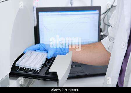 Un ricercatore che mette la piastra PCR sul termociclatore per l'amplificazione del DNA. Grafico delle curve sul monitor dietro. Test PCR del coronavirus. Foto Stock