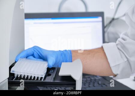 Un ricercatore che mette la piastra PCR sul termociclatore per l'amplificazione del DNA. Grafico delle curve sul monitor dietro. Test PCR del coronavirus. Foto Stock