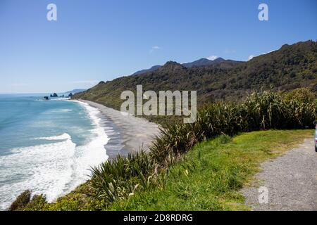 Una spiaggia di mare nella costa occidentale della Nuova Zelanda circondata da montagne e cespugli verdi e alberi Foto Stock